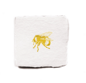 Pictured is a piece of handmade paper in a rough square. It has a gold letterpressed bee in the center of it. This is a Bee Petite Charm by Oblation sold at The Hare & The Hart.