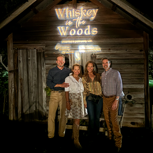 Pictured are four people standing in front of a sign that reads "Whiskey in the Woods."