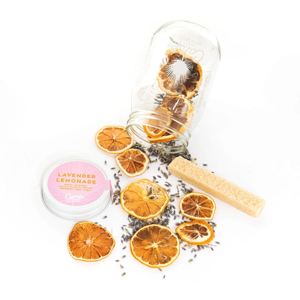 Pictured is a Lavender Lemonade Camp Craft Cocktail being sold at The Hare & The Hart in Thomasville, GA. The glass mason jar containing the kit is tipped over and the lid is off. The contents of the kit are spilled onto a plain white surface. The ingredients include dried lavender and dried orange slices.