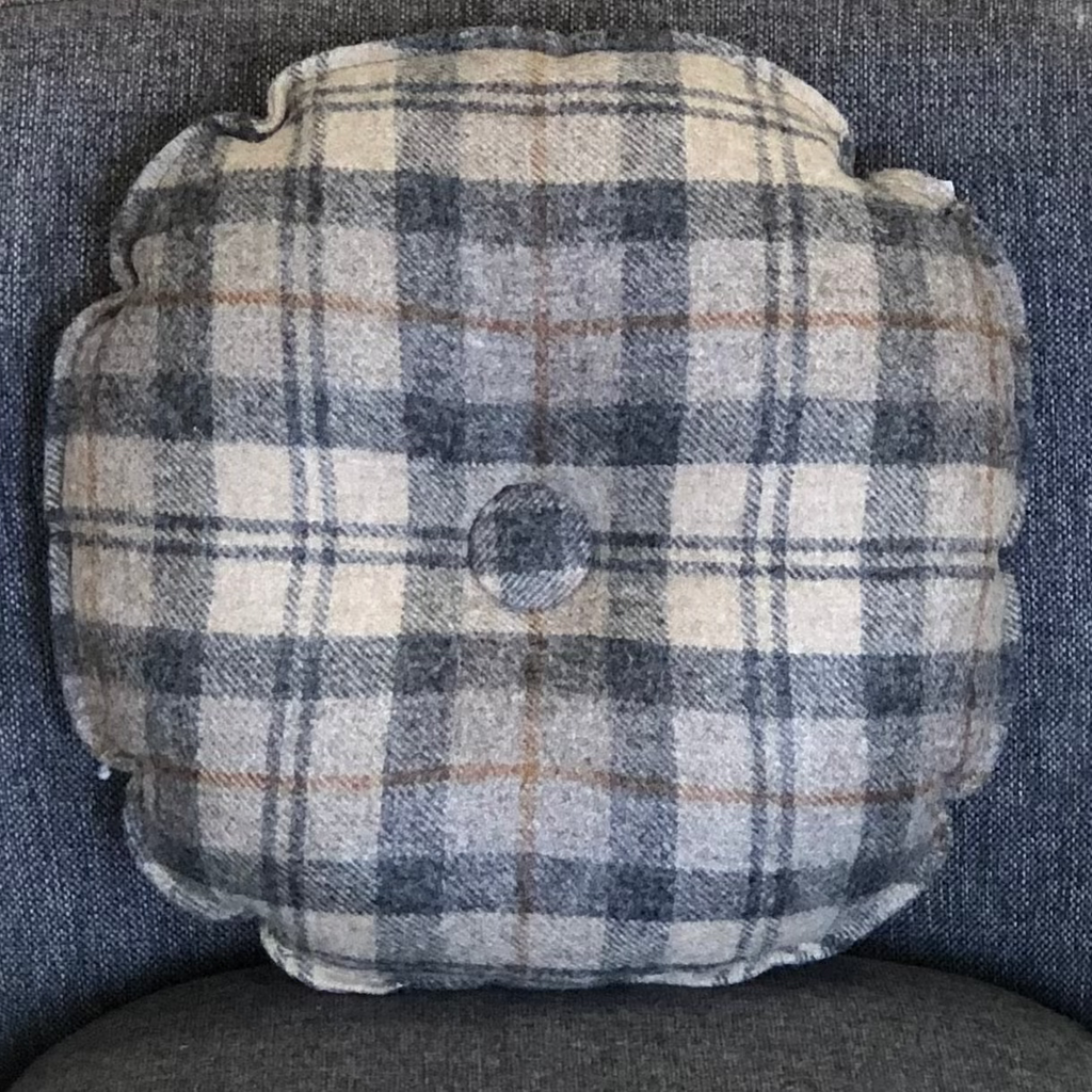 Pictured is Wool Throw Pillow Plaid 17” Round Pillow with Button by Honey Bee Fabrications sold at the gift shop The Hare & The Hart in downtown Thomasville, Georgia. It is a round throw pillow covered in gray plaid fabric.