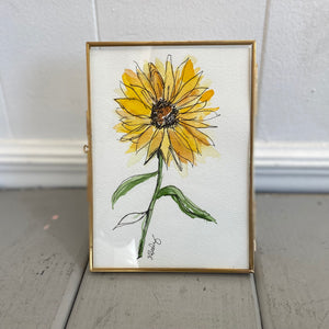 Pictured is Sunflower 5" x 7" Painting by Sarah Healy sold at The Hare & The Hart in downtown Thomasville, Georgia. It is a watercolor painting of a sunflower with pen and ink details in a thin gold frame.