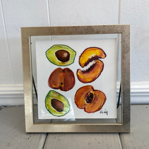 Pictured is Peaches & Avocados 8x8 Painting by Sarah Healy sold at The Hare & The Hart in downtown Thomasville, Georgia. It is a painting of peaches and avocados in a gold frame.