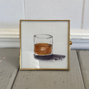Pictured is Bourbon Glass 4" x 4" Painting by Sarah Healy sold at The Hare & The Hart in downtown Thomasville, Georgia. Pictured is a watercolor painting of a glass of bourbon in a thin gold frame.