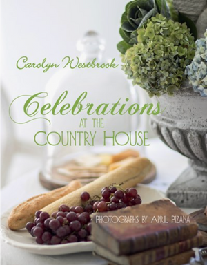 Pictured is the book "Celebrations at the Country House" by Carolyn Westbrook with photographs by April Pizana sold at The Hare & The Hart in Thomasville, GA. On the cover of the book is a plate with bread and grapes sitting next to a pile of antique books and a concrete vase filled with blue and green hydrangeas.
