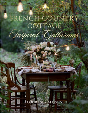 Pictured is the book "French Country Cottage Inspired Gatherings" by Courtney Allison sold at The Hare & The Hart in Thoamsville, GA. On the cover is a ornately set dining table sitting in a garden with string lights hung above it. Centered on the table is an unusually large bouquet of white roses and greenery. The chairs around the table are mismatched and the setting is equally haphazard, but in a cozy and sophisticated way.