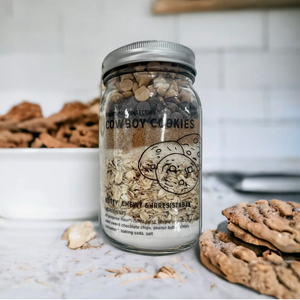 Pictured is a jar of Cowboy Cookie Mix in 32-oz Jar sold at The Hare & The Hart in downtown Thomasville, Georgia. It is next to a plate of baked cookies.