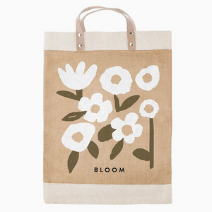 Pictured is Bloom Tote Bag sold at The Hare & The Hart in downtown Thomasville, Georgia. It is a natural tote bag printed with white flowers and green leaves and short handles.