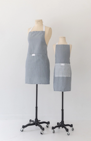 Pictured is an old fashioned mannequin wearing a Keepsake Jar Inen Adult Bistro Apron in Light Blue by Heirloomed Collection sold at The Hare & The Hart in Thomasville, Georgia. There are two mannequins, both wearing aprons, next to each other in the picture. One mannequin wears an adult sized apron and the other wears at child sized apron. The adult apron is light blue in color with white straps and one large pocket on the front center.