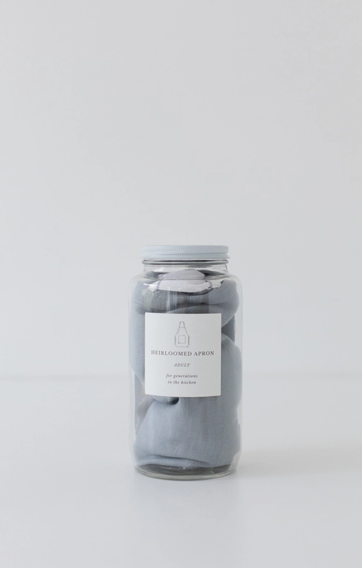 Pictured is a glass jar containing the Keepsake Jar Linen Adult Bistro Apron in Light Blue by Heirloomed Collection. The apron is inside of a clear glass jar with a white lid.