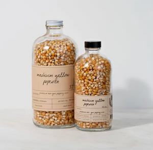Pictured is Medium Traditional Yellow Popcorn by Stone Hollow Farmstead sold at The Hare & The Hart in Thomasville, Georgia. There are two glass bottles next to each other, one 16 oz. and the other 32 oz. Each has a label on it describing the popcorn. They are each visibly filled with popcorn kernels.