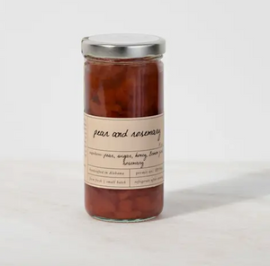 Pictured is Pear and Rosemary Preserves by Stone Hollow Farmstead sold at The Hare & The Hart in Thomasville, Georgia. The preserves are in a clear glass jar with a metal lid and a tan label describing them. The preserves inside are a deep red color.