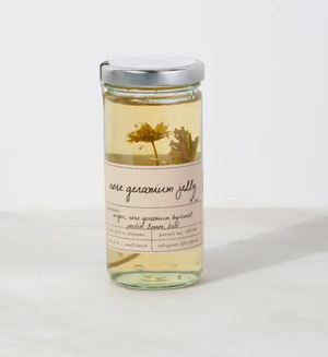 Pictured is Rose Geranium Floral Jelly by Stone Hollow Farmstead sold at The Hare & The Hart in Thomasville, Georgia. It is in a clear glass jar with a metal lid and a tan label. Inside is a yellow tinted clear jelly with two preserved flowers suspended inside.