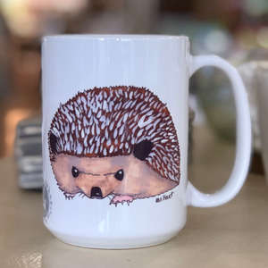 Pictured is a white coffee mug with a watercolor design of a hedgehog on it.