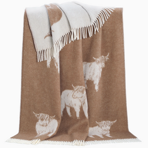 Pictured is a Highland Cow Wool Throw by J. J. Textiles sold at The Hare & The Hart in downtown Thomasville, Georgia. It is a brown and white blanket with highland cow designs on it. It has tassels.