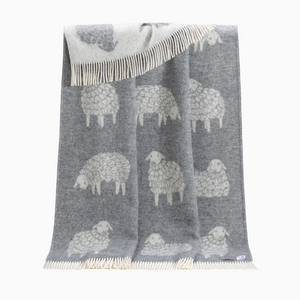 Pictured is a Sheep Wool Throw in Grey by J. J. Textile sold at The Hare & The Hart in downtown Thomasville, Georgia. It is a wool blanket in grey and white with sheep designs on it. It has tassels.