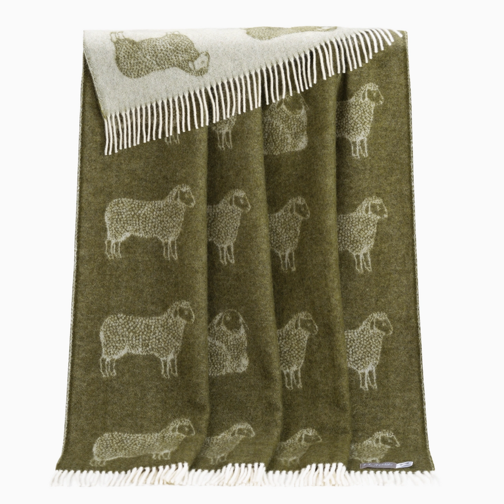 Pictured is a Sheep Wool Throw by J.J. Textile in Green sold at The Hare & The Hart in downtown Thomasville, Georgia. It is a green and white blanket with images of sheep on it. It has tassels.