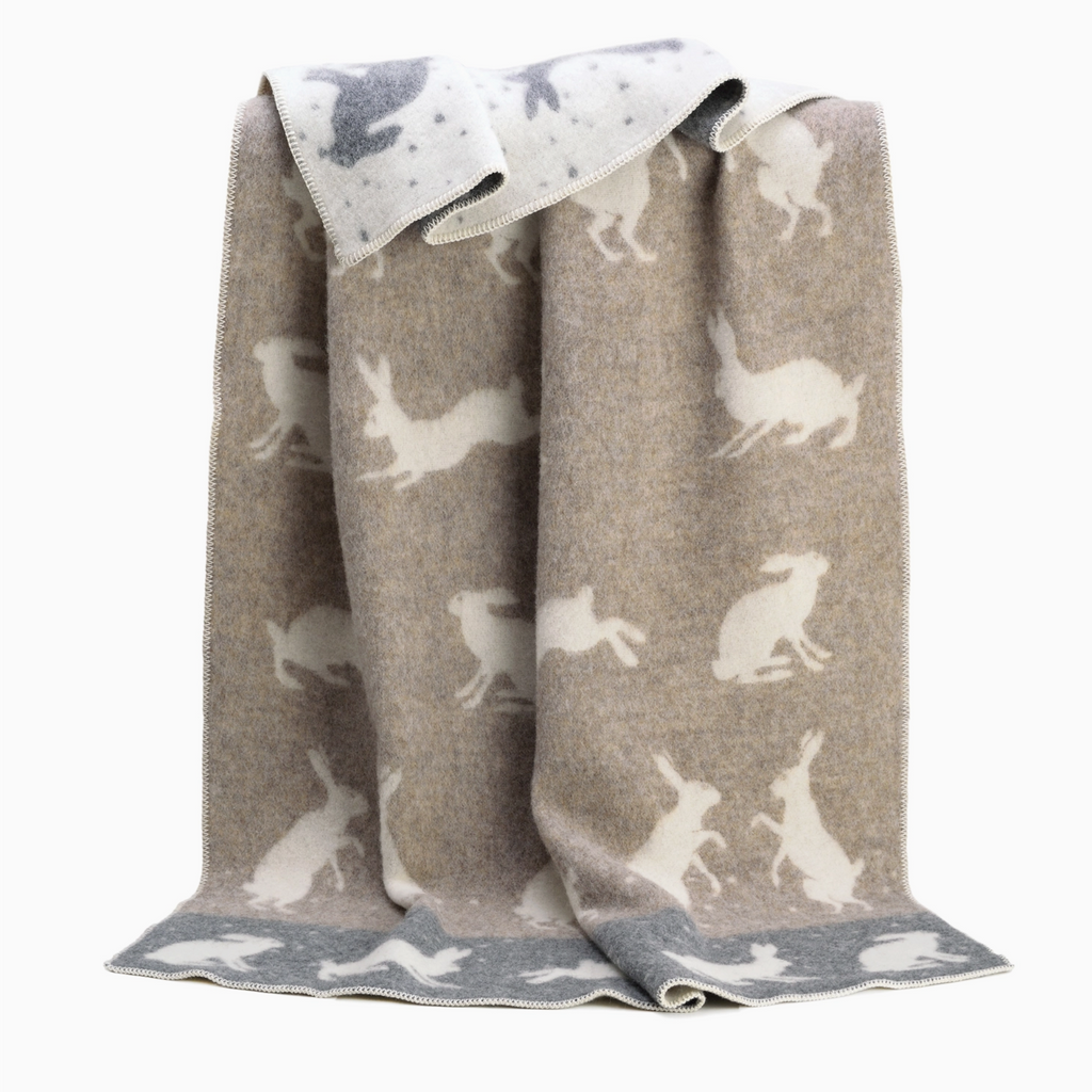 Pictured is a Boxing Hares Wool Blanket by J.J. Textile sold at The Hare & The Hart in downtown Thomasville, Georgia. It is a blanket with rabbit designs on it. It is gray and white.
