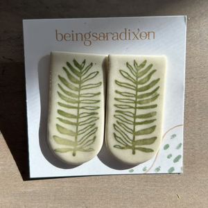 Pictured is Fern Porcelain Statement Stud Earrings sold at The Hare & The Hart in downtown Thomasville, Georgia. They are large ceramic stud earrings with green ferns painted onto them.
