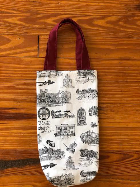 Pictured is a fabric wine tote. The body of the bag is made of Toile of FSU black and white fabric. The two handles are made of garnet fabric. The tote lays on a wood floor.