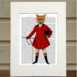 A page from a book is framed with mat. Printed over the page is a figure wearing an old hunting outfit with a red coat. In their left hand is the crop and tucked under their right arm is their riding helmet. Instead of the head of a person they have the head of a fox. This image is a full body portrait of the fox figure. This is a Fox Hunter Book Print by Fab Funky Ltd. sold at The Hare & The Hart.