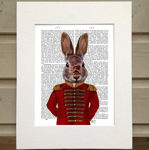 Pictured is a page from a book framed with mat. Printed over the top of the page is a figure in an old military coat. The coat is red with gold details. The figure has the head of a cute rabbit rather than the head of a person. The rabbit is pictured from the waist up and its arms are tucked behind its back.