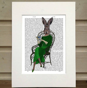 Pictured is a page from a book framed with mat. Printed over top the text on the book page is a figure sitting in a metal chair. The chair is organic in its style. The figure wears a quarter sleeve, full lenght green dress and a pair of matching pointed shoes. The figure holds a teacup and a small plate. Instead of the head of a person the figure has the head of a hare with its ears alert.