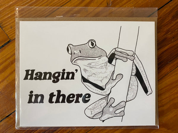 There is a horizontal greeting card with a white backgrounda and a drawing of a frong hanigng from a branch in black. It has the words "Hangin' in there" printed on it in a serif font. This is a Notecard by Birds & Bees sold at The Hare & The Hart.