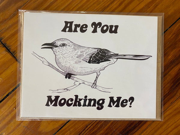 There is a horiztonal greeting card. The card has a whtie background with the design of a mocking bird on it. There are also the words "Are You Mocking Me?" This is a Notecard by Birds & Bees sold at The Hare & The Hart.
