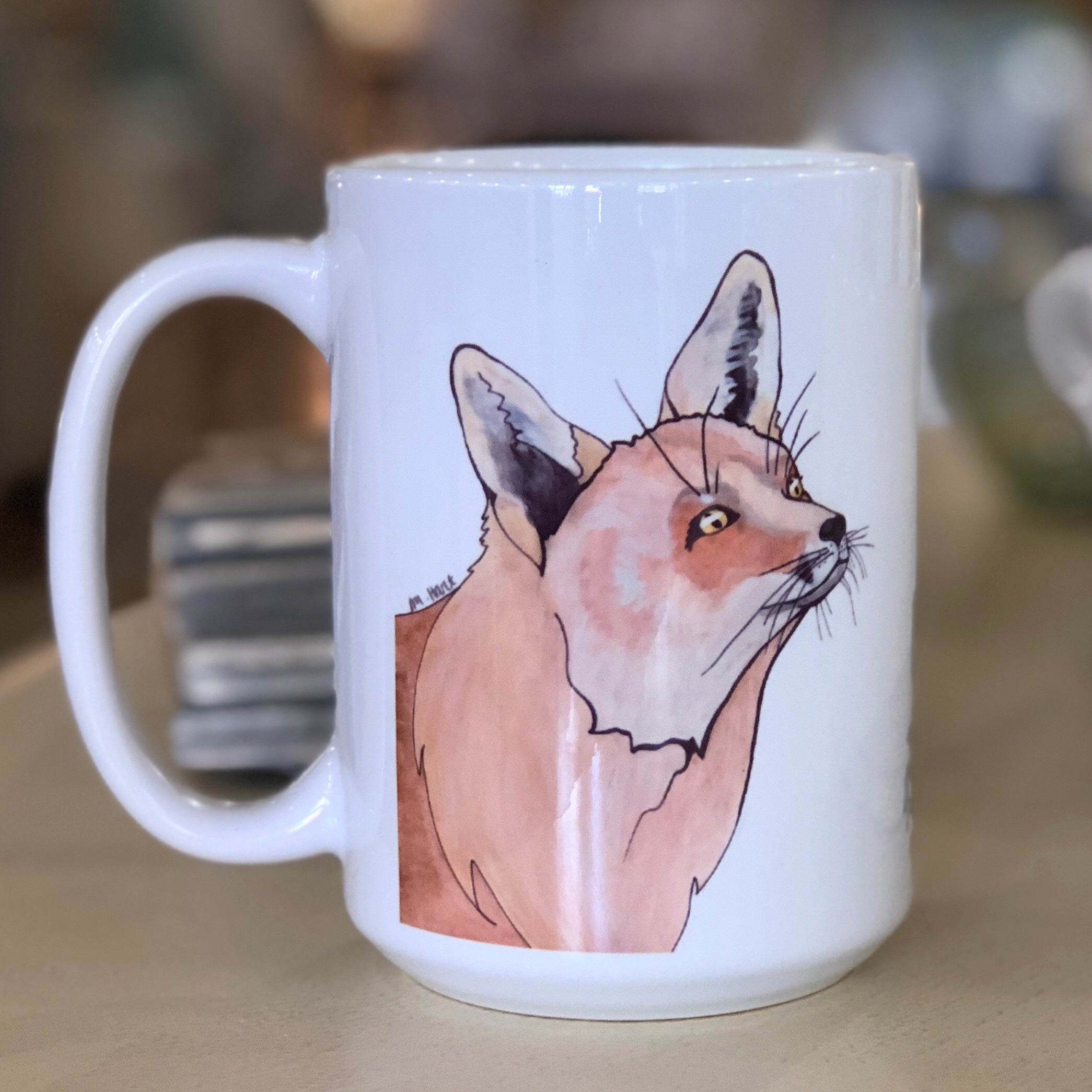 Pictured is a white coffe mug with a watercolor design of a fox on it.