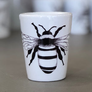 A shot glass is pictured. It has a pure white background with one black bee design in the center. This is a Bee Shot Glass by Birds & Bees sold at The Hare & The Hart.