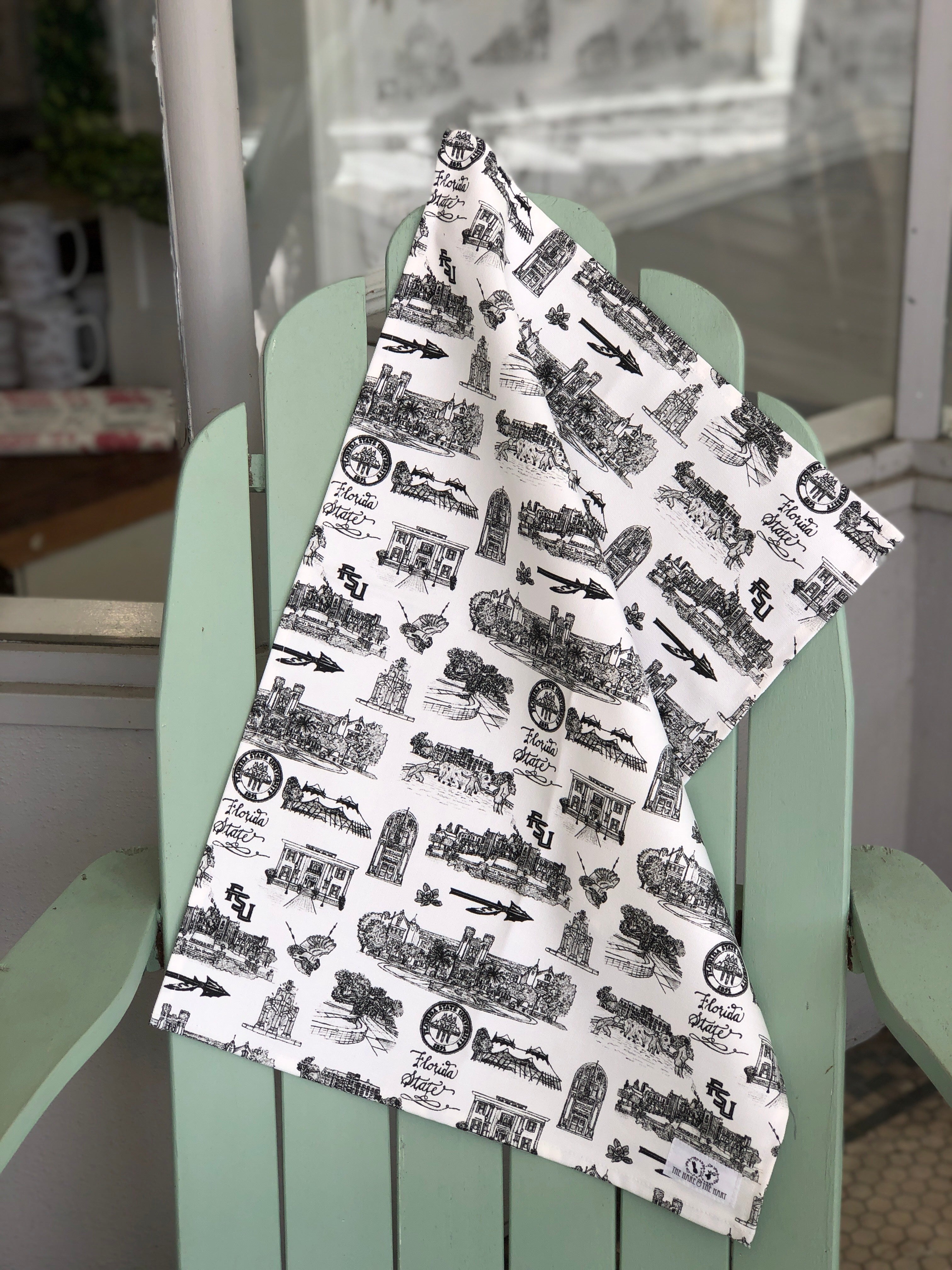 There is a tea towel made of Toile of FSU patterned fabric in black and white. The tea towel is hanging from the top of an adirondak chair.