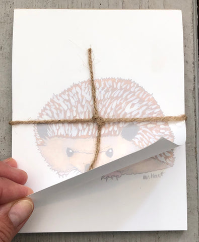 There is a notepad tied together with twice. The paper of the notepad is printed with a design of a hedgehog. The first page of the notepad is being held open by a hand.