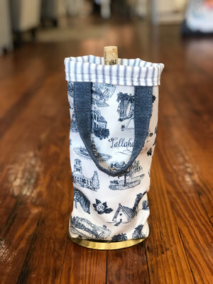 There is a blue and white Toile of Tallahassee wine bag containing a bottle of wine. The top edge of the bag is folded over, showing the blue and white ticking stripe fabric lining.