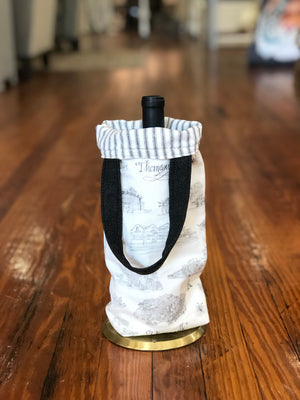 Pictured is a black and white Toile of Thomasville wine bag with a bottle of wine inside. The handles are black and the lining inside is black and white ticking stripe.