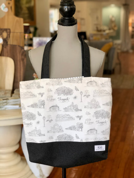 There is a black and white Toile of Thomasville tote bag hanging from the neck of a mannequin. The bottom of the bag and the handles are made of black fabric.