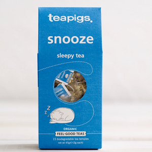 Pictured is the packaging for Teapigs snooze tea. It is a rectangular blue box with an illustration of a sleeping cat on it. There is a circular clear plastic window through which the teabags can be seen. The label reads "teapigs snooze sleepy tea organic feel-good teas."