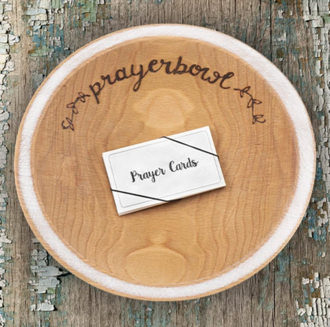 Pictured is the Grace Prayer Bowl. It is carved out of wood and has a natural finish. The lip of the bowl is wide and painted white. Carved into the bowl along one edge, in cursive, is the words "prayerbowl" and two flower designs on either side. Inside the bowl is a stack of prayer cards.