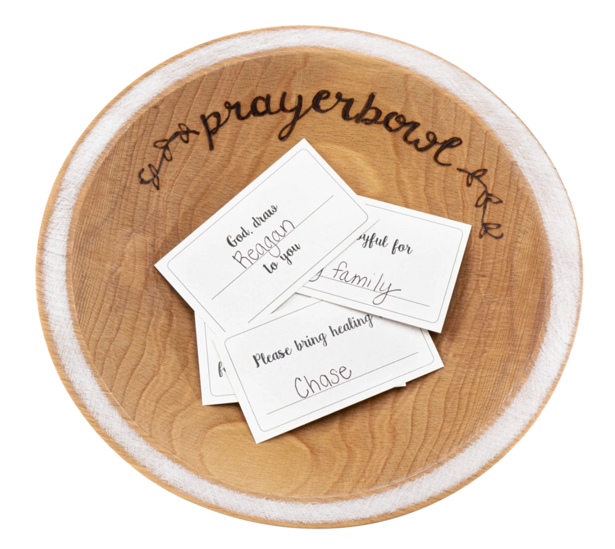 Pictured is the Grace Prayer Bowl. It is carved out of wood and has a natural finish. The lip of the bowl is wide and painted white. Carved into the bowl along one edge, in cursive, is the words "prayerbowl" and two flower designs on either side. Inside the bowl is a pile of prayer cards.