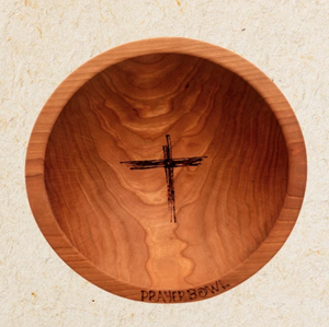 Pictured is the Noah Prayer Bowl. It is carved from wood and has a warm finish. The shape of the bowl is very simple. There is a rough carving of a cross in the center of the bowl's basin. On the bottom edge lip of the bowl are carved the words "PRAYER BOWL."