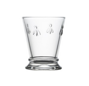 A clear glass mini tumbler with four embossed bees on opposite sides. It is in the shape of a tumbler glass but is shorter. This is a Bee Mini Tumbler by La Rochere sold at The Hare & The Hart.