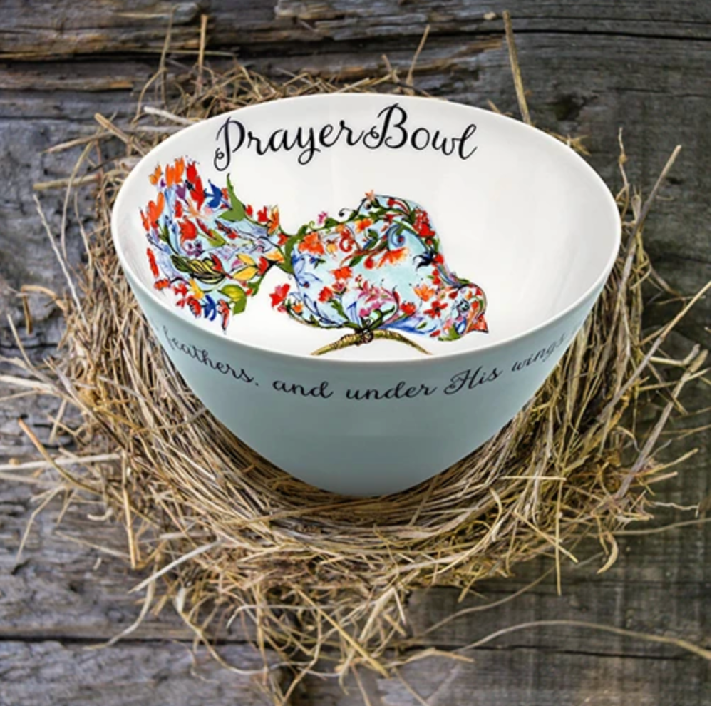 The Josephine Prayer Bowl is pictured from the side, so the painting inside is visible. The bowl is round and deep. Inside, it is painted white with the deisgn on a bird made of flowers perched on a limb taking up one wall of the bowl. Above the bird, in cursive script, are the words "Prayer Bowl." The outside of the bowl is light blue wth a border of cursive script.