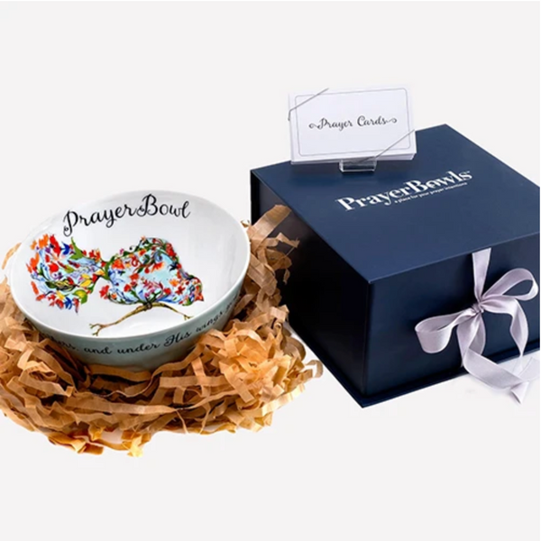 Pictured is the Jospehine Prayer Bowl nestled in strips of decorative brown paper, like a bird in a nest. Next to it is the dark blue box packaging it comes in and the prayer cards that accompany it.