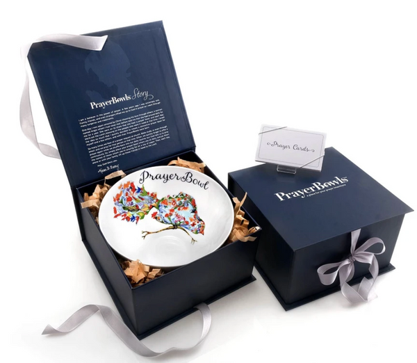 Pictured are two dark blue boxes used to package the Josephine Prayer Bowl. One is open, showing the prayer bowl inside, cushioned in decorative paper. The inner lid of the box has the Prayer Bowls story printed on it. The second box is unopened and its ribbon is tied in a bow.