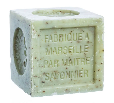 Pictured is a verbena soap block by Savon de Marseille. It is a light green cube of soap with brown flecks in it. Etched into the side of it are the words "FABRIQUE A MARSEILLE PAR MAITRE SAVONNIER."