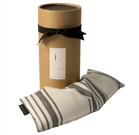 Pictured is a Lavande Eye Pillow next to its container. The container is a cylinder with a black ribbon around it and a white sticker label. The Eye Pillow itself is long and rectangular with gray and white stripes.