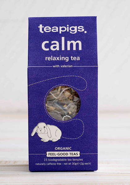 Pictured is the packaging for Teapigs Organic Calm Tea. It is a rectangular box that is dark blue and has the image of a white bunny on it. There is a circle of clear plastic to show the teabags inside the packaging. The label of the package reads "teapigs calm relaxing tea with variation organic feel-good teas."