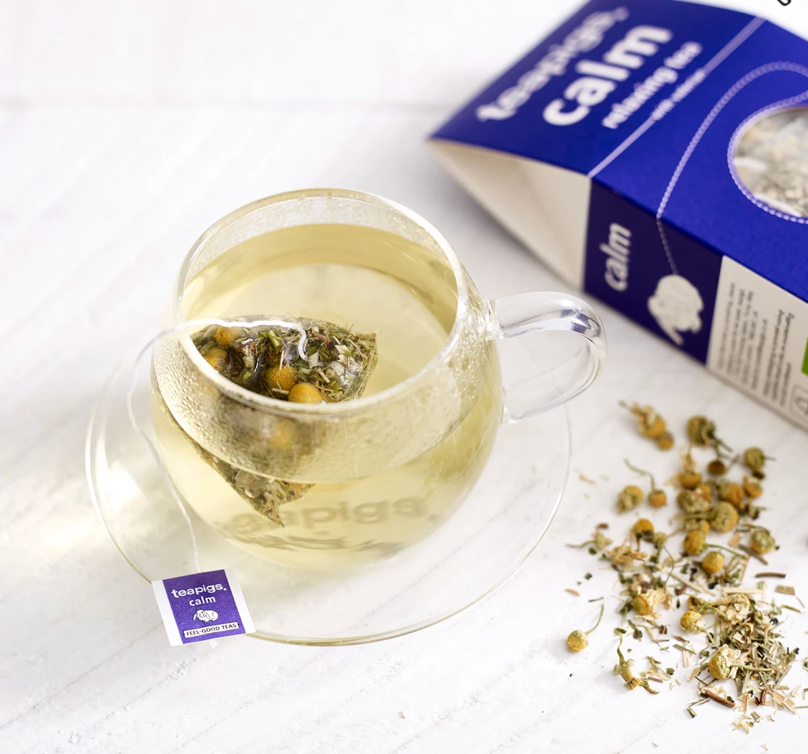 Pictured is a cup of tea next to loose tea and an open package teapigs organic calm tea.