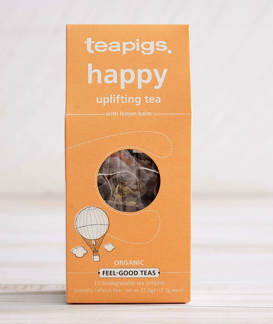Pictured is the packaging for Teapigs Organic Happy Tea. The box is a light orange color and has a design of a tiny hot air balllon. There is a small round clear plastic window through which the teabags can be seen. The label on the packing reads "teapigs happy uplifting tea with lemon balm organic feel-good teas"