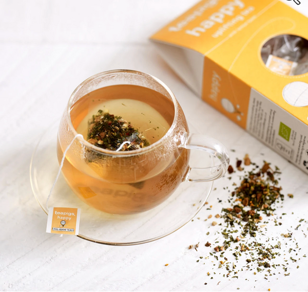 Pictured is a cup of tea next to loose leaf tea and an open package of teapigs organic happy tea.