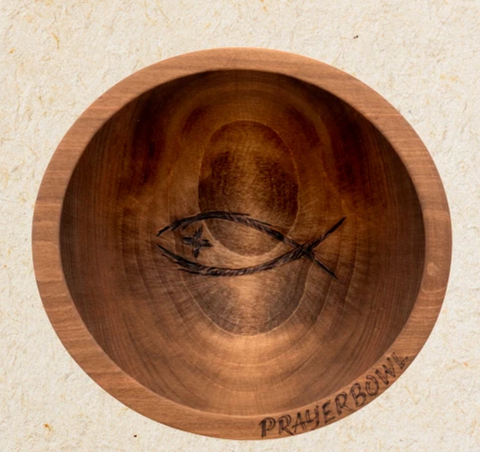 Pictured is the Randolph Prayer Bowl. It is a simple, shallow bowl carved from wood. In the center is a carving of an ichthys with a cross for an eye. On the bottom edge of the bowl is a carving of the words "PRAYER BOWL"